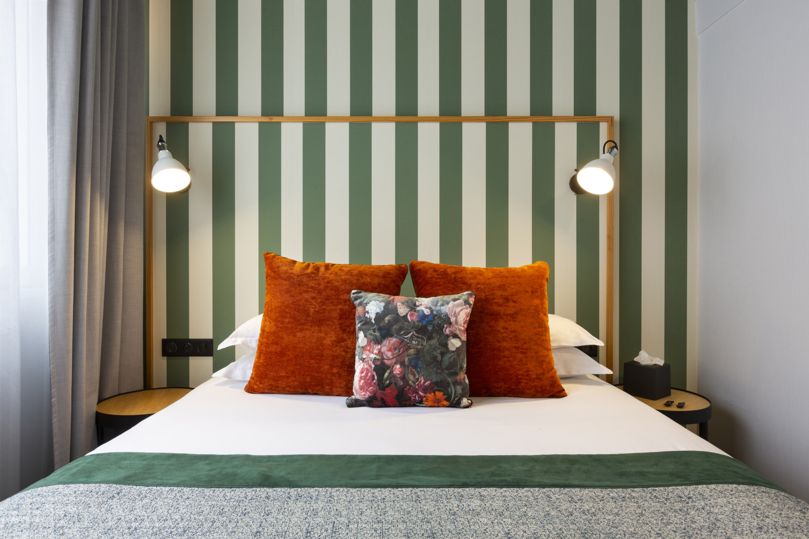 Doppelzimmer: Bett mit orangefarbenen und gebl&uuml;mten Kissen vor gr&uuml;n-wei&szlig; gestreifter Wand, zwischen zwei Wandleuchten und runden Nachttischen